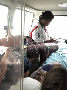 Ylva ligger i sykebilen og blir passet på av en representant fra Toten folkehøgskole og en lokal sykepleier. Bildet er tatt gjennom luken fra forsetet i bilen.