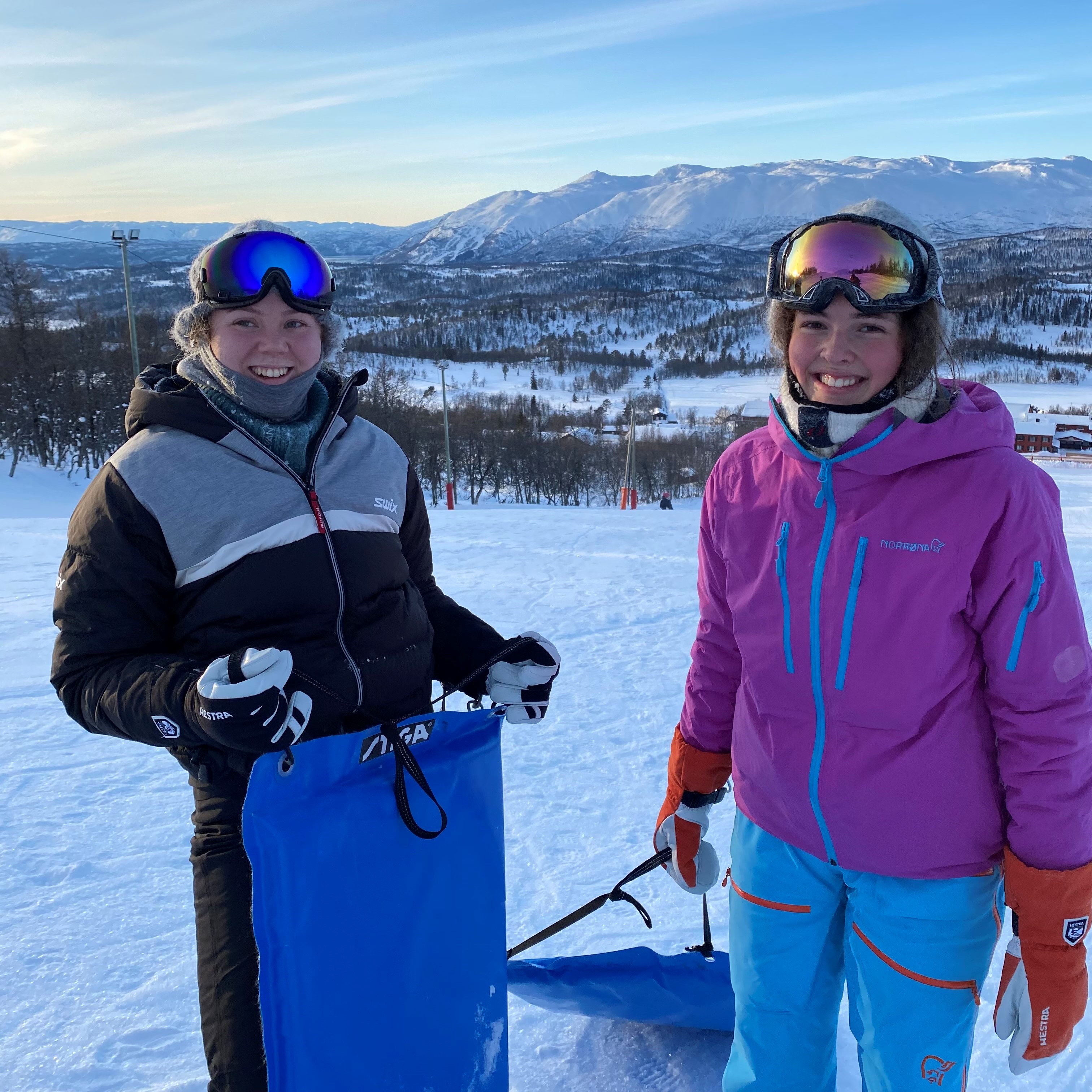 Nathalie og medelev i akebakken. Smiler. Fjell i bakgrunnen.