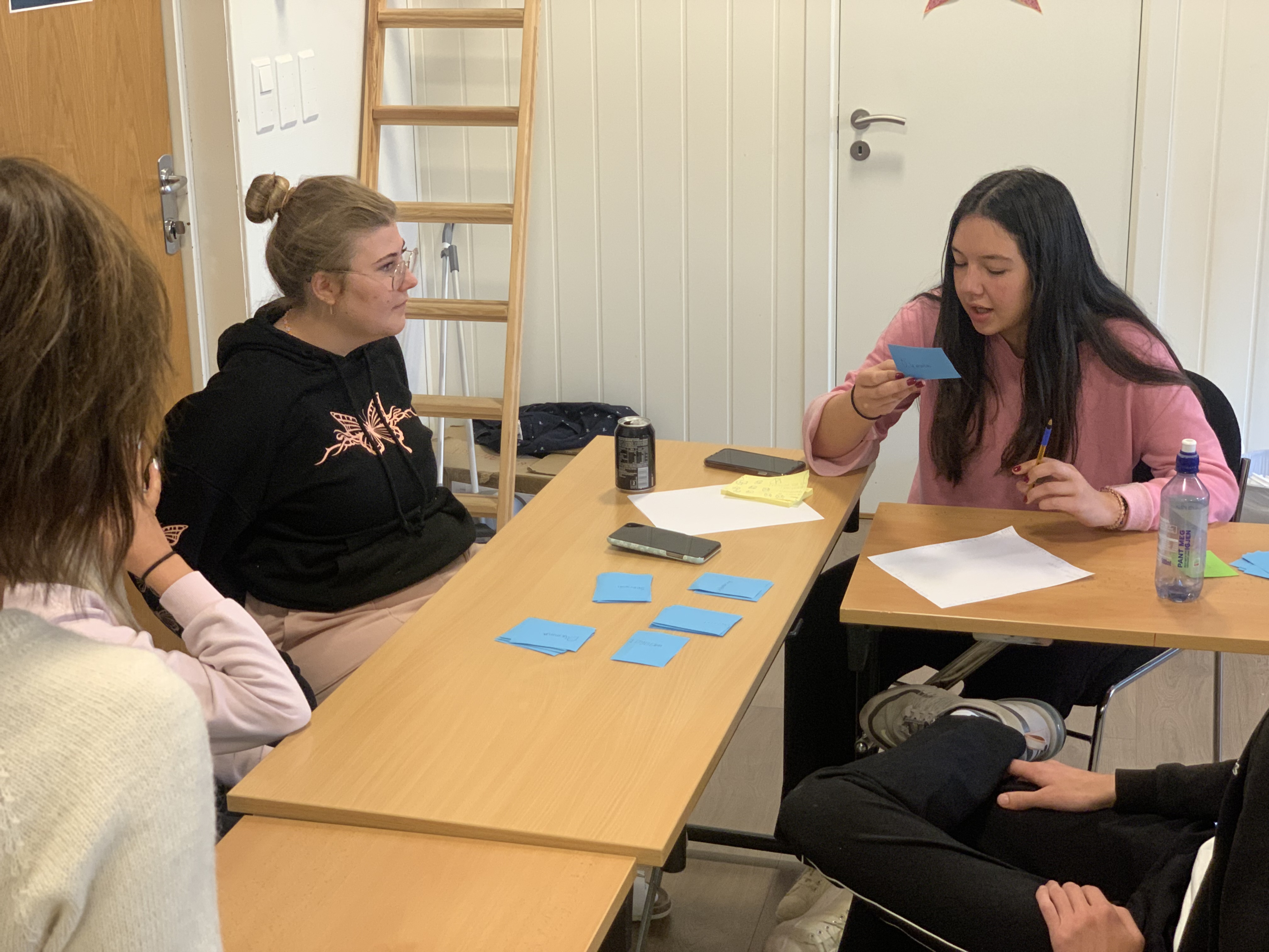 klasserom, tre elever. Mange post-it lapper på bordet, Sara leser opp fra en av dem.
