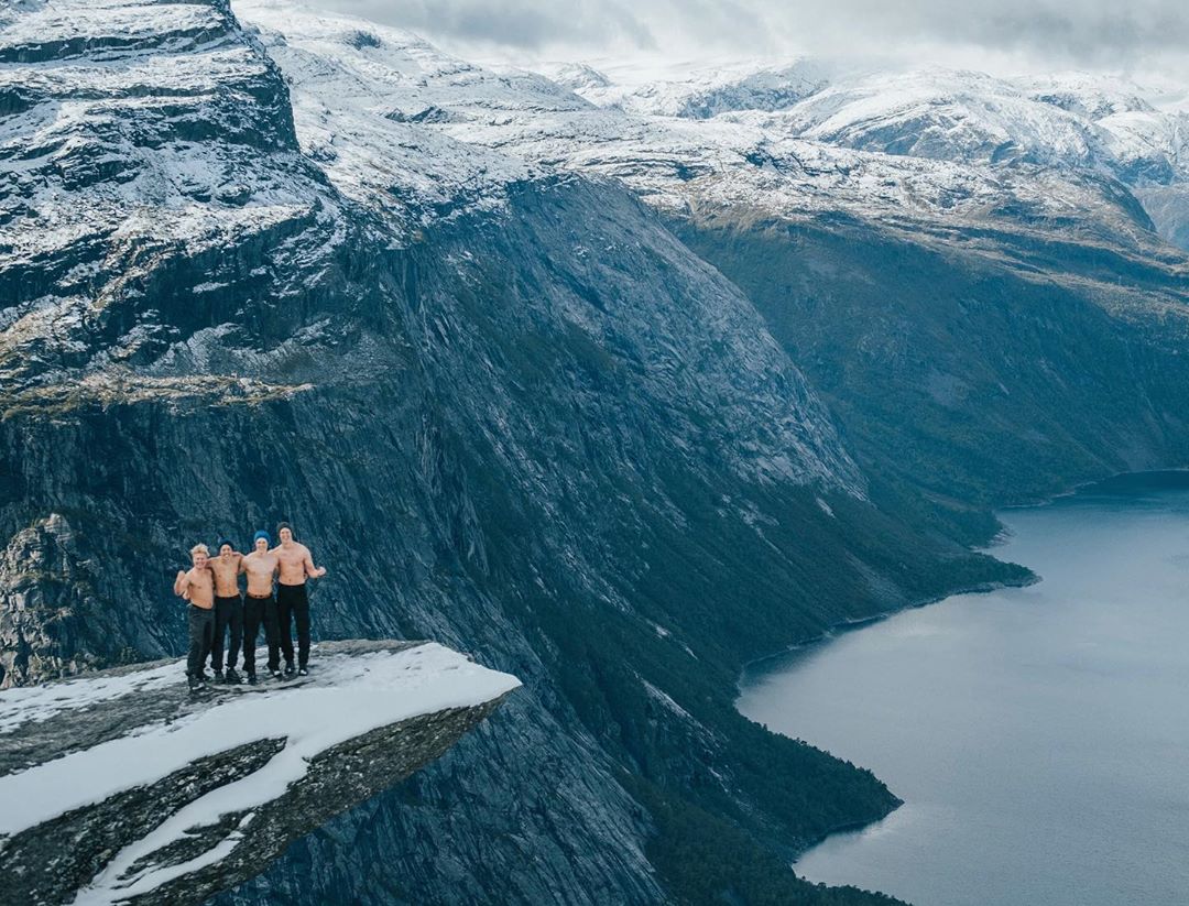 Fire personer med bar overkropp står på et fjellutsprang med utsikt til fjell og fjord.