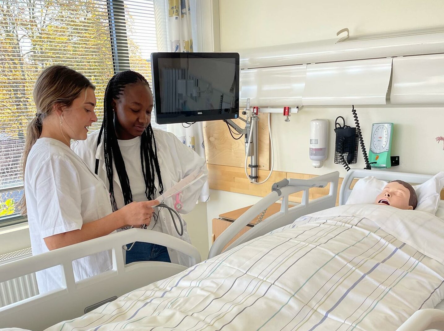 To ungdommer kledt i hvitt ser på en sykehusseng med en undervisningsdukke