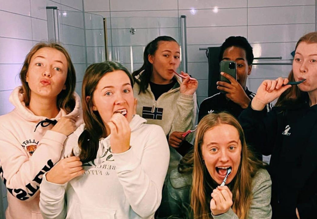 Flere jenter pusser tennene mens de ser i et speil. De er kledd i joggeklær og er usminka.