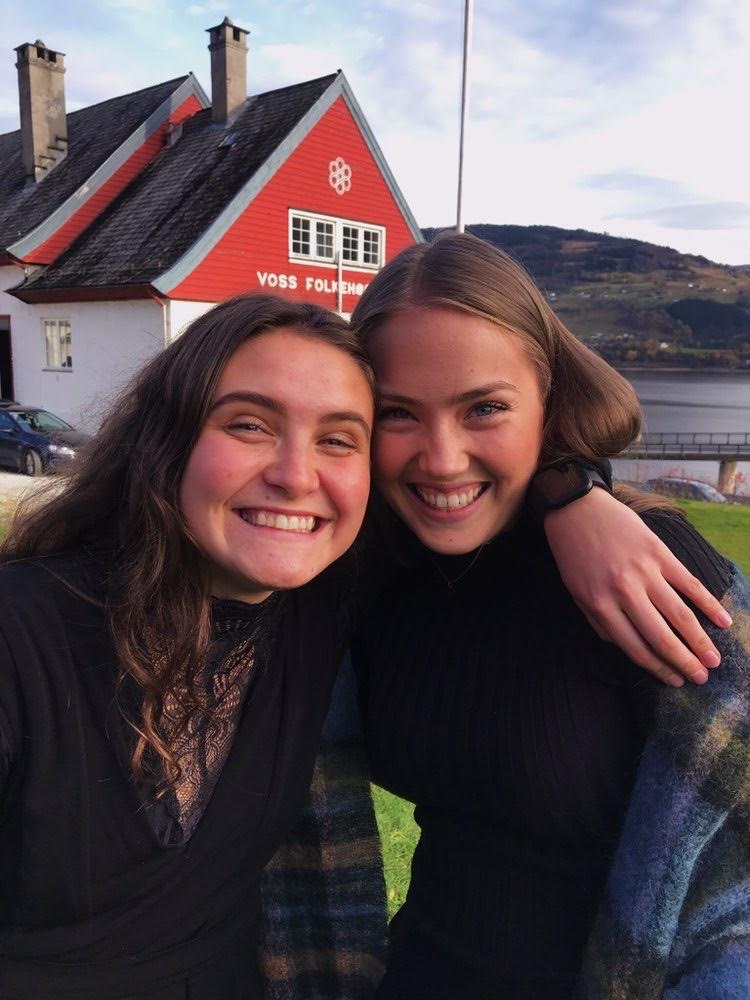 To smilende jenter klemmer foran et bygg med skiltet "Voss folkehøgskule"