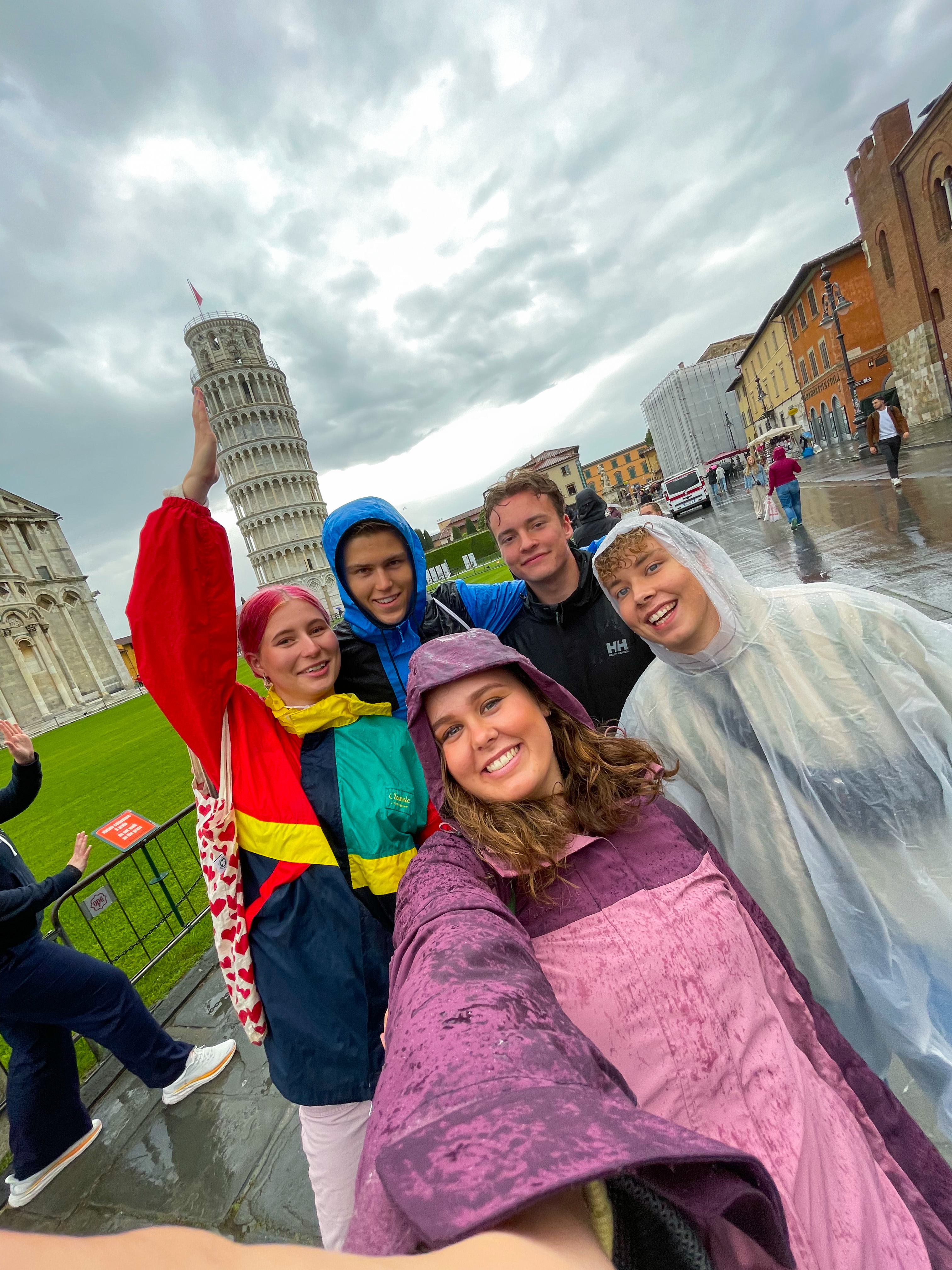 En gruppe unge mennesker i regntøy står foran det skjeve tårnet i Pisa
