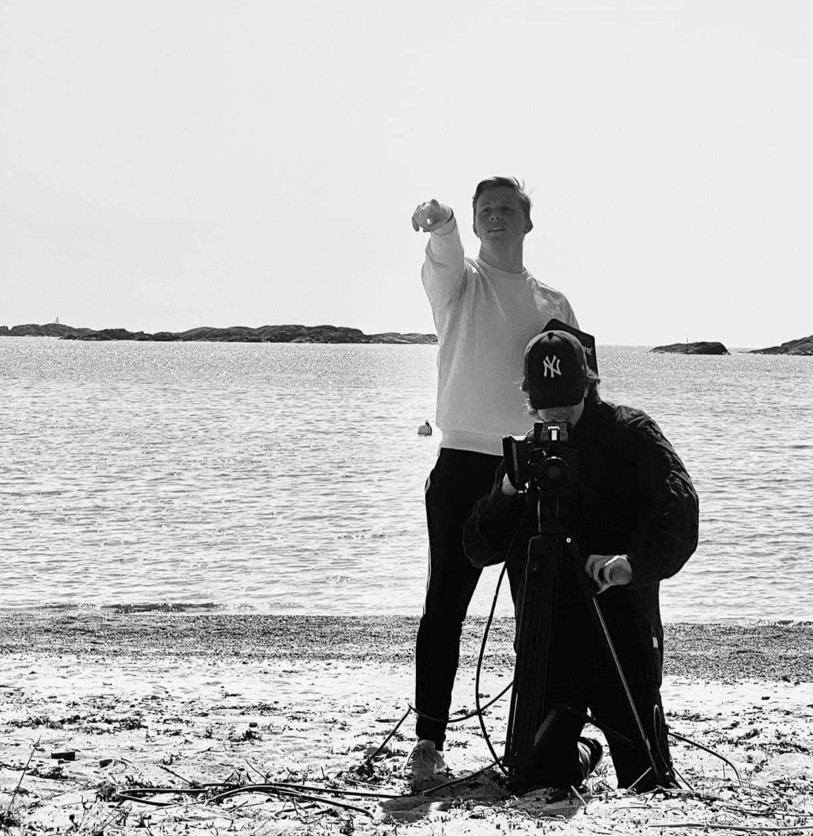 Mathias og medelev med filmkamera på en strand. Vannet i bakgrunnen