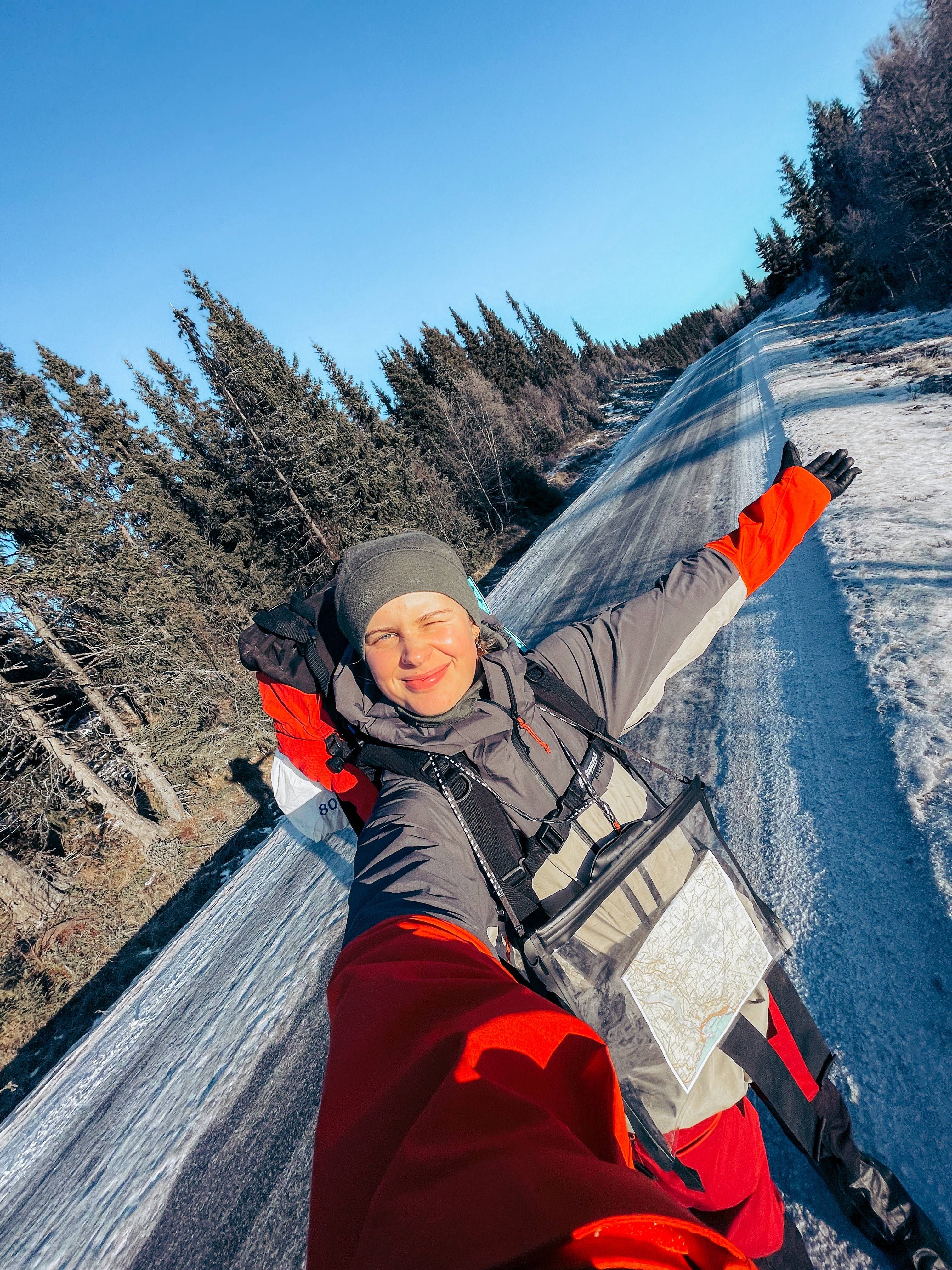 En kvinnelig elev tar selfie på tur om vinteren med sekk på ryggen og kartlomme på brystet.