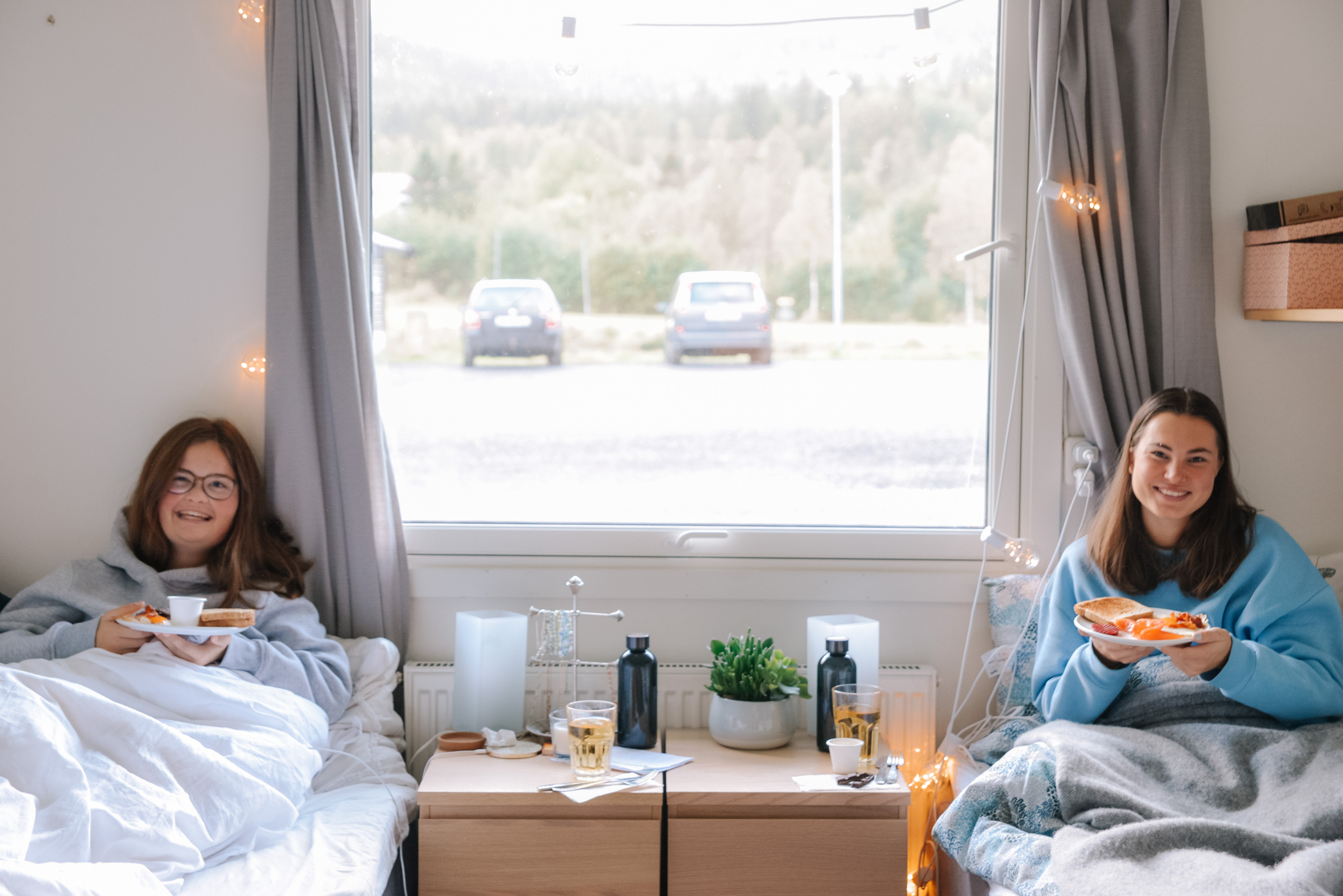 To unge kvinner ligger i hver sin seng og spiser frokost på senga.