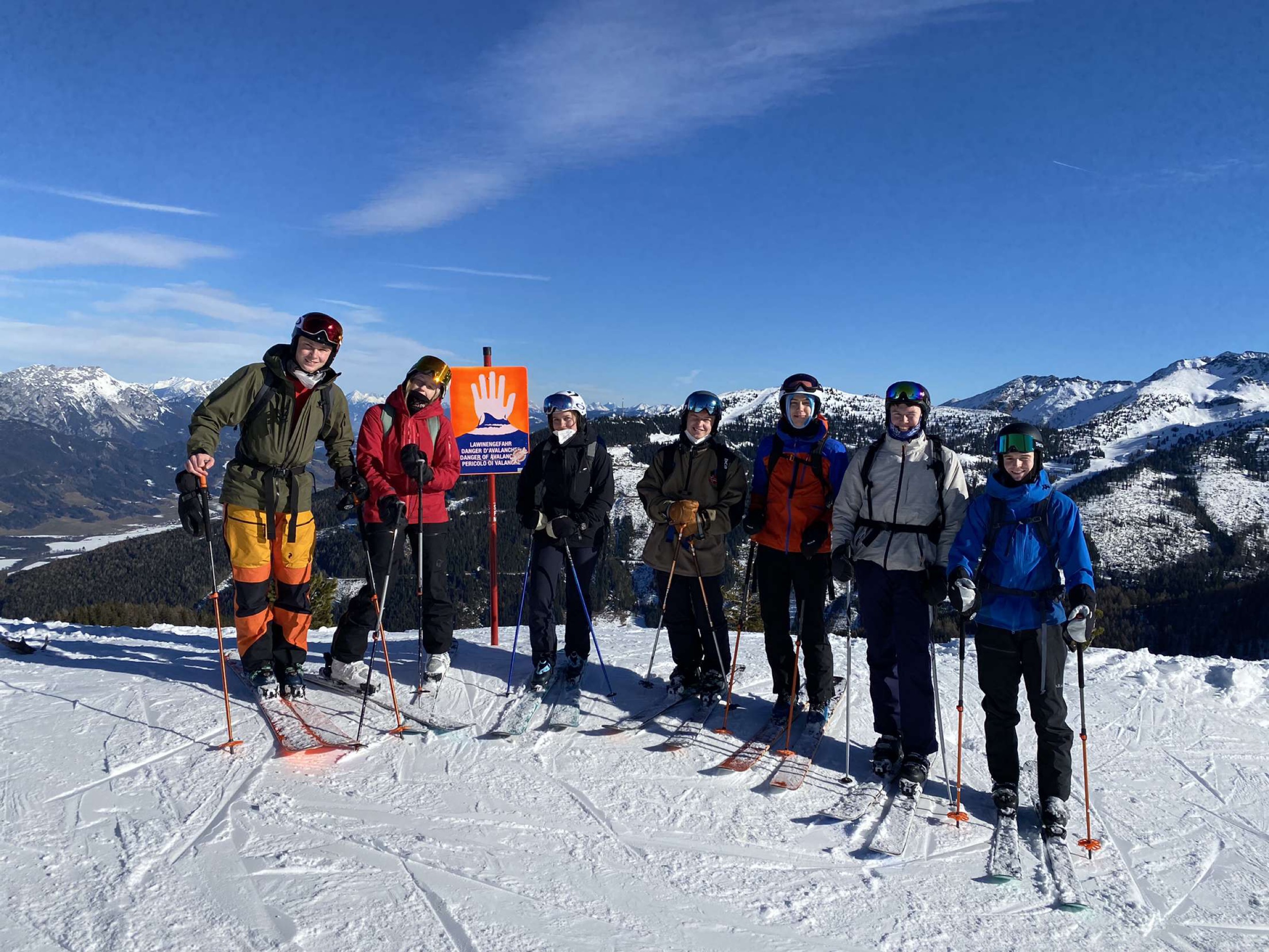 Snødekt bakke og glade ungdommer på ski. Blå himmel og fjell i bakgrunnen.