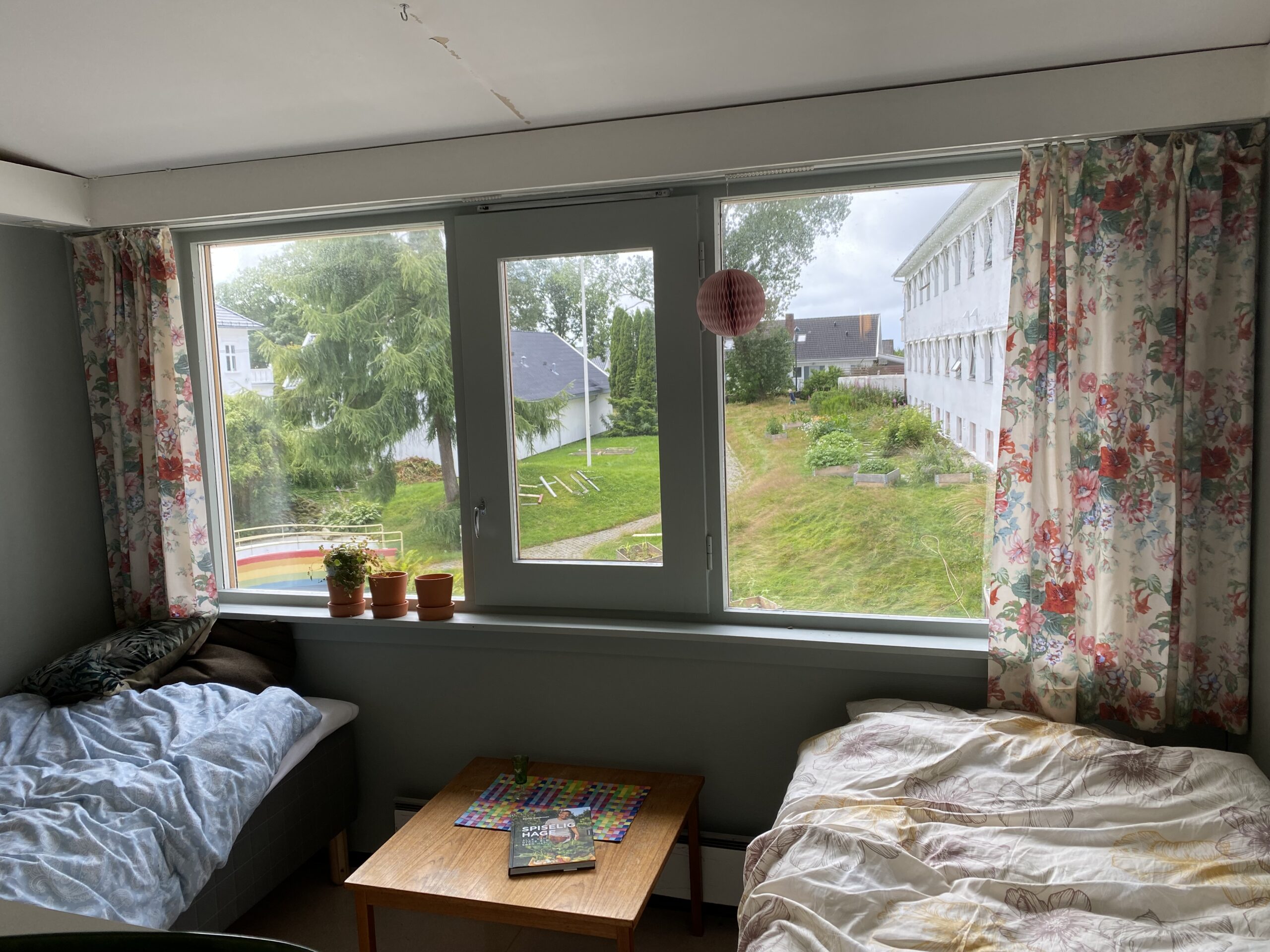 Bilde av rom med to senger og et vindu imellom sengene. Gjennom vinduet ser man en hage med en bro med en regnbue malt på siden