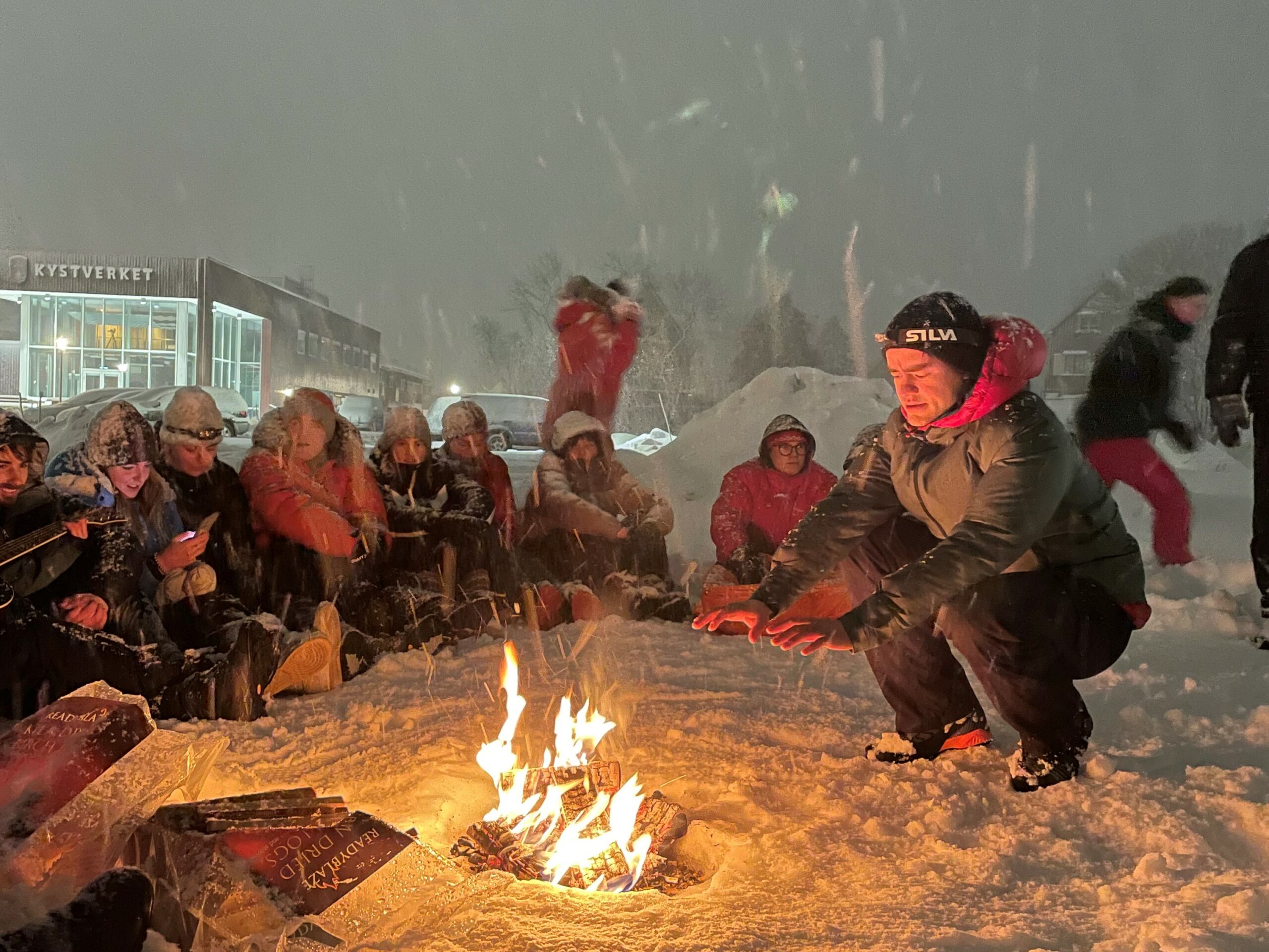 en mann som sitter på huk foran et bål sammen med en gruppe mennesker.