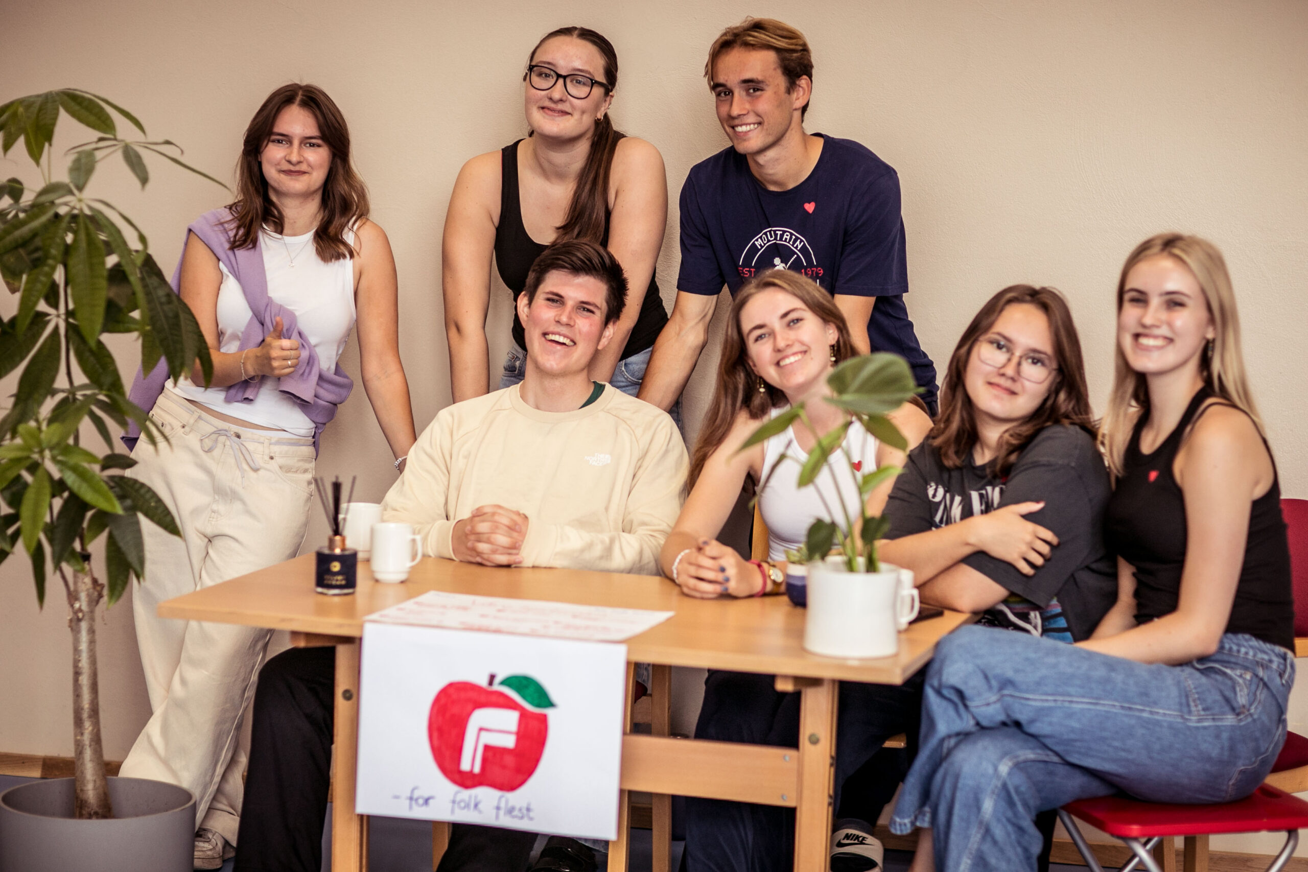 En gruppe ungdommer sitter og står rundt et bord med et skilt med logoen til Fremskrittspartiet