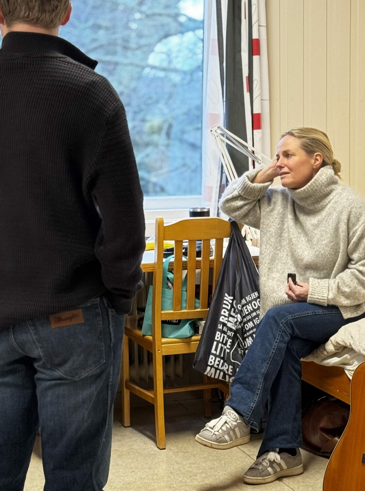 en kvinne som sitter på en seng på et internatrom, hun snakker med en ung mann med ryggen til kamera