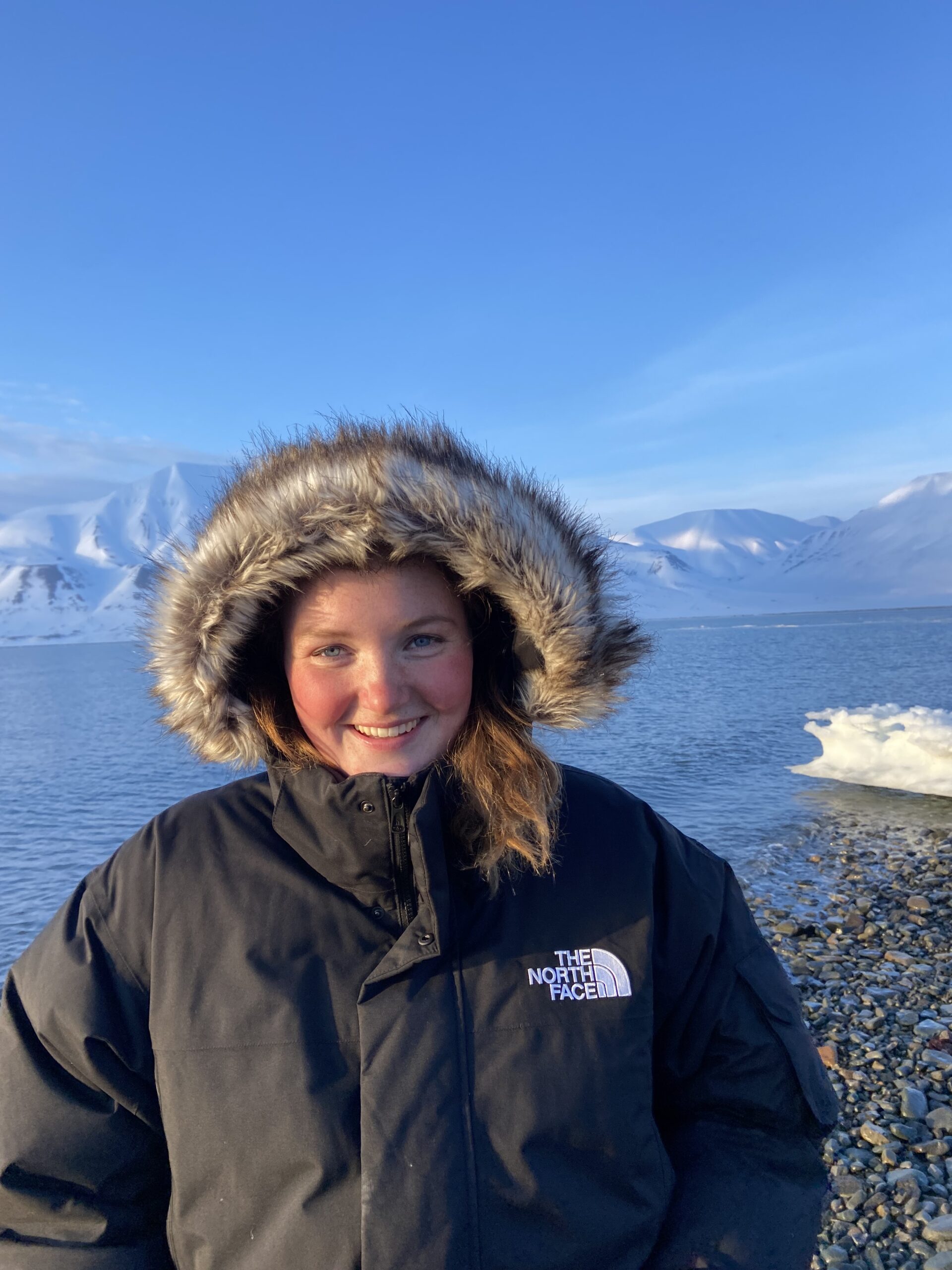 En jente smiler i kamera i idylliske svalbardomgivelser i pelshette