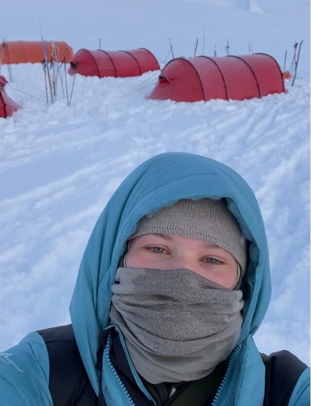 Karoline står i snø med telt bak seg, fullt påkledd i buff og hette
